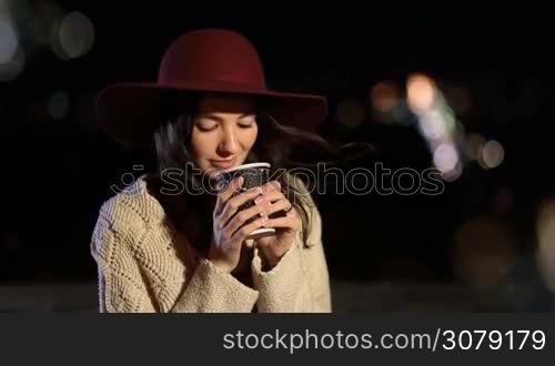Fashionable young woman with long brown hair sitting on the bench in park at night and drinking hot fresh coffee from paper cup over colorful streetlights background. Attractive female enjoying night city landscape with coffee to go.