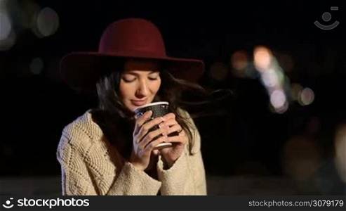 Fashionable young woman with long brown hair sitting on the bench in park at night and drinking hot fresh coffee from paper cup over colorful streetlights background. Attractive female enjoying night city landscape with coffee to go.