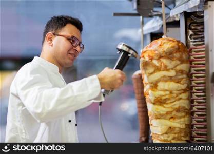 fast food and cooking concept - chef slicing doner meat from rotating spit at kebab shop. chef slicing doner meat from spit at kebab shop