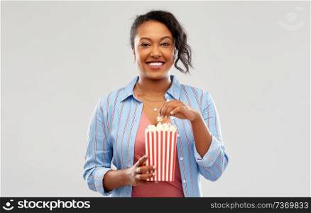 fast food and people concept - happy african american young woman eating popcorn from striped bucket over grey background. happy african american woman eating popcorn
