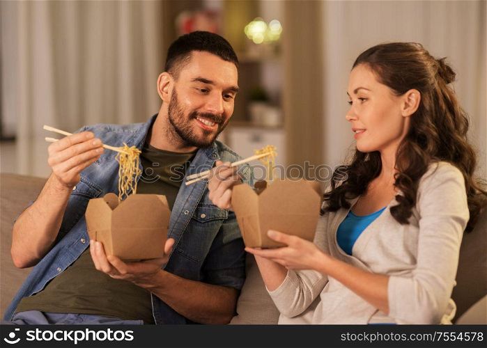 fast food and people concept - happy couple eating takeaway noodles with chopstick at home in evening. happy couple eating takeaway noodles at home