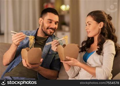 fast food and people concept - happy couple eating takeaway noodles with chopstick at home in evening. happy couple eating takeaway noodles at home
