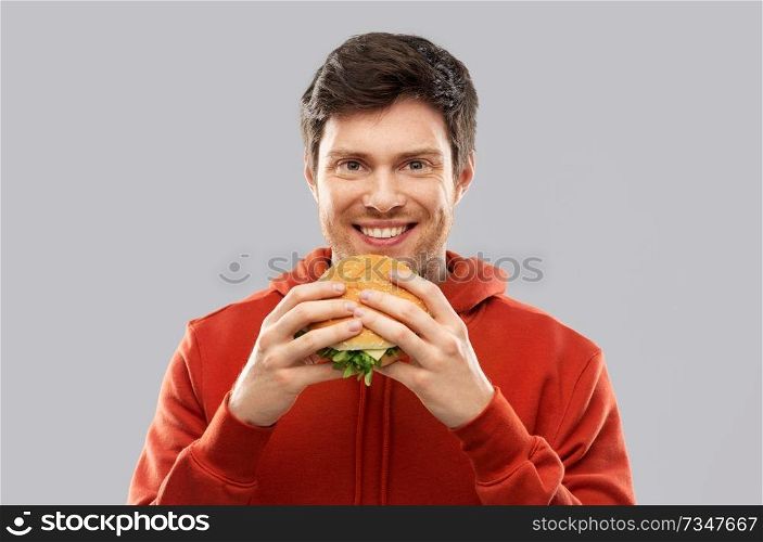 fast food and people concept - happy smiling young man in red hoodie eating hamburger over grey background. happy young man eating hamburger