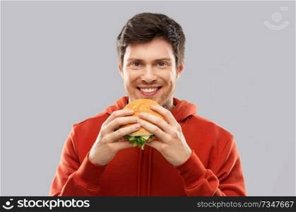 fast food and people concept - happy smiling young man in red hoodie eating hamburger over grey background. happy young man eating hamburger