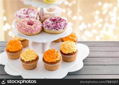 fast food and sweets concept - glazed donuts and cupcakes with buttercream frosting on stand over festive lights background. glazed donuts, cupcakes with frosting on stand