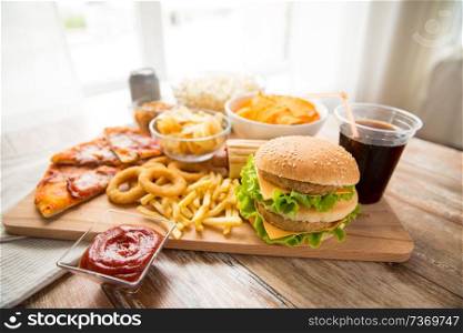 fast food and unhealthy eating concept - close up of double hamburger or cheeseburger, deep-fried squid rings, french fries, pizza and cola drink on wooden board. close up of fast food and cola drink on table