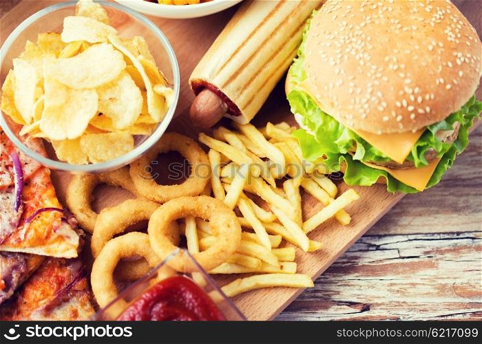 fast food and unhealthy eating concept - close up of hamburger or cheeseburger, deep-fried squid rings, french fries hotdog and potato chips on wooden table top view