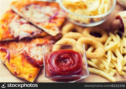 fast food and unhealthy eating concept - close up of ketchup in glass bowl over pizza, deep-fried squid rings, potato chips, peanuts and ketchup on wooden table