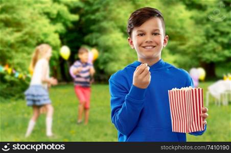 fast food, childhood and people concept - happy smiling boy in blue hoodie eating popcorn from striped paper bucket over friends having fun at birthday party in summer park background. smiling boy eating popcorn at birthday party