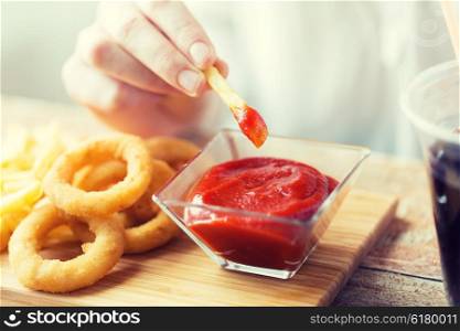 fast food, people and unhealthy eating concept - close up of hand with deep-fried squid rings, dipping french fries into ketchup bowl on wooden table