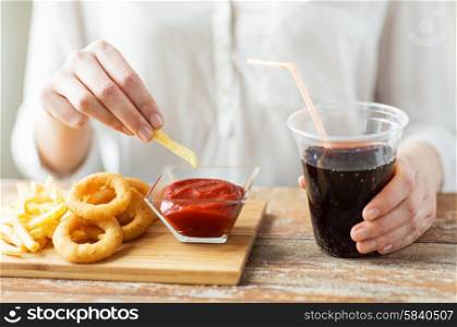 fast food, people and unhealthy eating concept - close up of woman with deep-fried squid rings, dipping french fries into ketchup bowl and drinking coca cola on wooden table