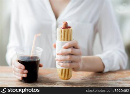 fast food, people and unhealthy eating concept - close up of woman hands with hot dog and coca cola drink sitting at table