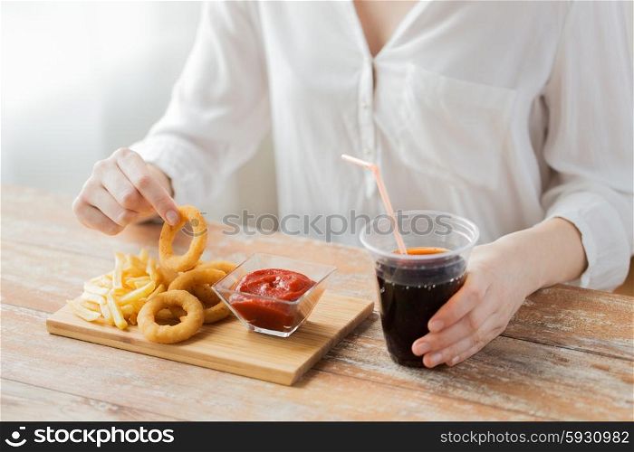 fast food, people and unhealthy eating concept - close up of woman eating deep-fried squid rings, french fries with ketchup and drinking coca cola on wooden table