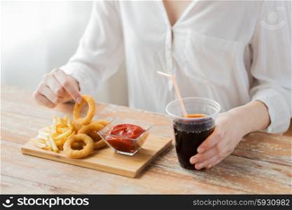 fast food, people and unhealthy eating concept - close up of woman eating deep-fried squid rings, french fries with ketchup and drinking coca cola on wooden table