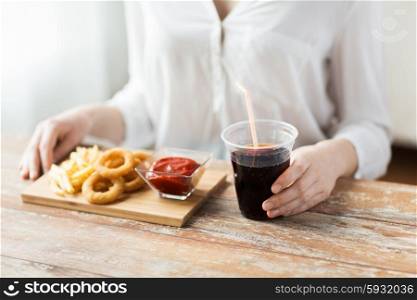 fast food, people and unhealthy eating concept - close up of woman eating deep-fried squid rings, french fries with ketchup and drinking cola on wooden table