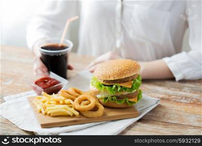 fast food, people and unhealthy eating concept - close up of woman hands with hamburger or cheeseburger, french fries, squid rings and cola drink sitting at table