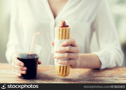 fast food, people and unhealthy eating concept - close up of woman hands with hot dog and cola drink sitting at table