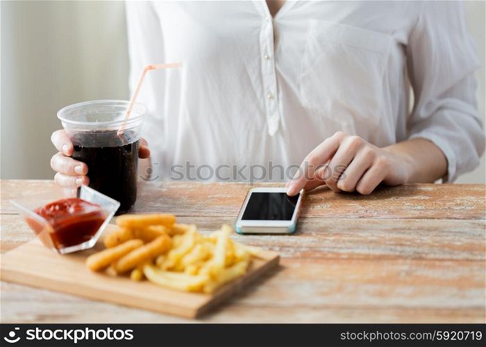 fast food, people, technology and diet concept - close up of woman with smartphone drinking coca cola and eating french fries, ketchup and deep-fried squid rings at wooden table