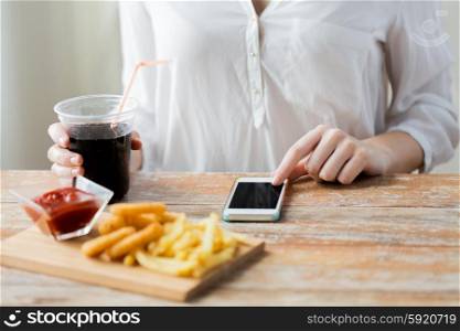 fast food, people, technology and diet concept - close up of woman with smartphone drinking coca cola and eating french fries, ketchup and deep-fried squid rings at wooden table