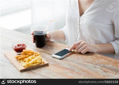 fast food, people, technology and diet concept - close up of woman with smartphone drinking cola and eating french fries, ketchup and deep-fried squid rings at wooden table