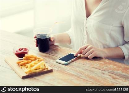 fast food, people, technology and diet concept - close up of woman with smartphone drinking cola and eating french fries, ketchup and deep-fried squid rings at wooden table