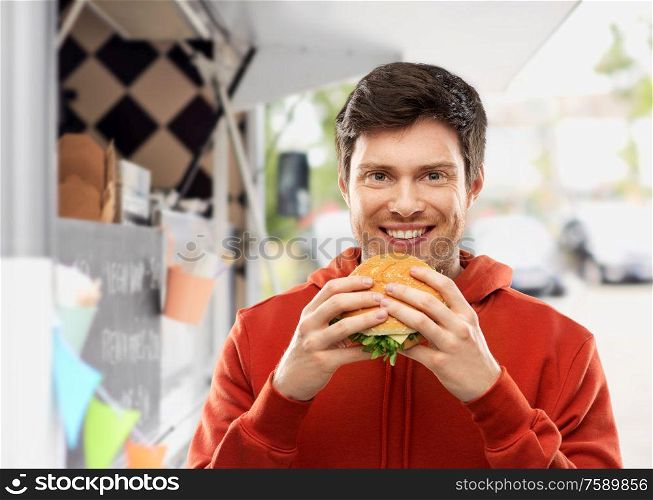 fast food, takeaway and people concept - happy smiling young man in red hoodie eating hamburger over food truck on street background. happy young man eating hamburger at food truck