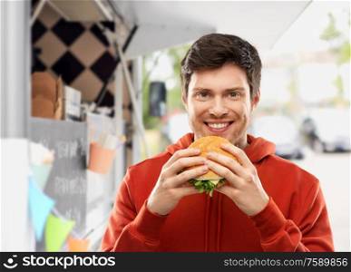 fast food, takeaway and people concept - happy smiling young man in red hoodie eating hamburger over food truck on street background. happy young man eating hamburger at food truck