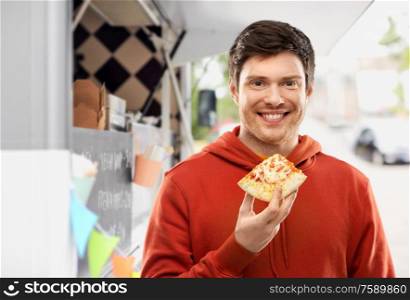 fast food, takeaway and people concept - happy smiling young man in red hoodie eating takeaway pizza over food truck on street background. happy young man eating pizza at food truck