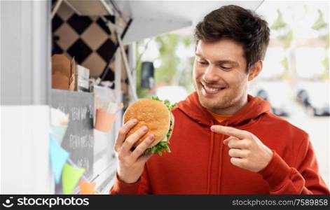 fast food, takeaway, eating and people concept - happy smiling young man in red hoodie showing hamburger over food truck on street background. happy young man showing hamburger at food truck