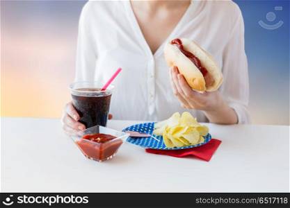 fast food, unhealthy eating and people concept - close up of woman with hot dog, cola in plastic cup, potato chips and ketchup over evening sky background. close up of woman eating hot dog with cola. close up of woman eating hot dog with cola
