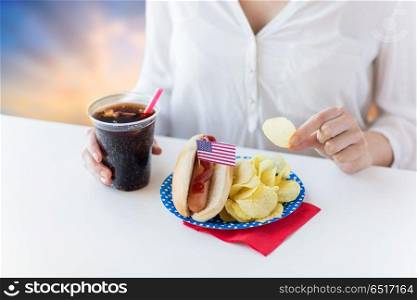 fast food, unhealthy eating and people concept - close up of woman with hot dog, cola in plastic cup and chips on independence day over evening sky background. close up of woman eating hot dog with cola. close up of woman eating hot dog with cola