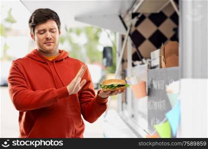 fast food, unhealthy eating and people concept - young man in red hoodie refusing from hamburger over food truck on street background. young man refusing from hamburger at food truck
