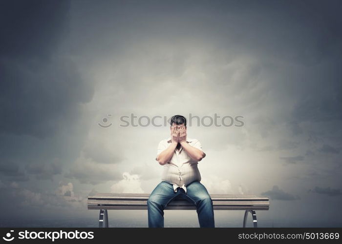 Fat man sitting on bench closing eyes with hands. Fat man