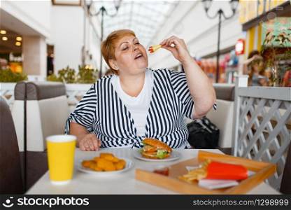 Fat woman eating burger in mall food court. Overweight female person at the table with junk lunch, obesity problem. Fat woman eating burger in mall food court