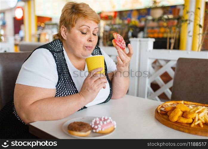 Fat woman eating doughnuts in fastfood restaurant, unhealthy food. Overweight female person at the table with junk dinner, obesity problem. Fat woman eating doughnuts in fastfood restaurant