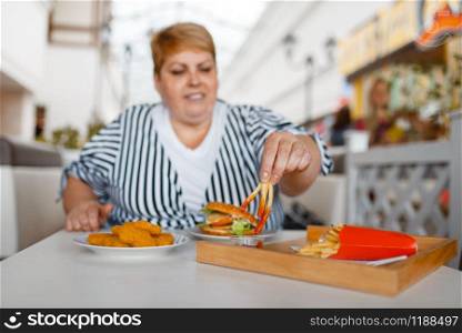Fat woman eating french fries in mall food court. Overweight female person at the table with junk lunch, obesity problem