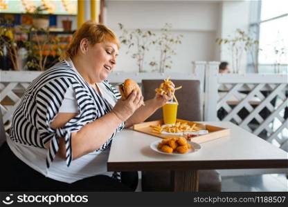 Fat woman eating high calorie food in fastfood restaurant. Overweight female person at the table with junk dinner, obesity problem. Fat woman eating high calorie food in restaurant