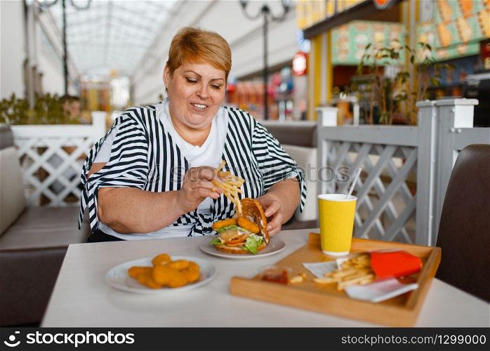 Fat woman eating high calorie food in mall restaurant. Overweight female person at the table with junk dinner, obesity problem. Fat woman eating high calorie food in mall