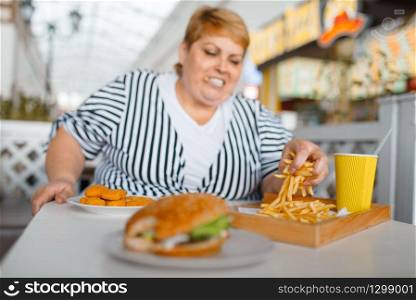 Fat woman eating high calorie food in mall restaurant. Overweight female person at the table with junk dinner, obesity problem