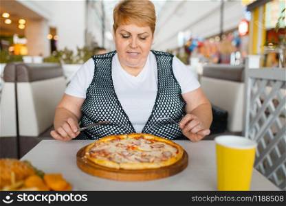 Fat woman eating pizza in fastfood restaurant. Overweight female person at the table with junk dinner, obesity problem