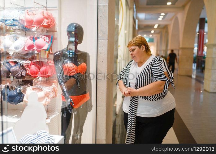 Fat woman near the showcase with underwear for slim body. Overweight female person dreaming at the store with lingerie, obesity problem. Fat woman near the showcase with underwear