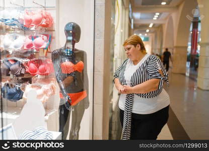 Fat woman near the showcase with underwear for slim body. Overweight female person dreaming at the store with lingerie, obesity problem. Fat woman near the showcase with underwear