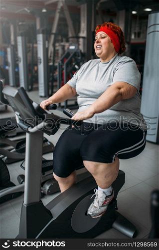 Fat woman training on exercise bike in gym. Calories burning, obese female person in sport club. Fat woman training on exercise bike in gym
