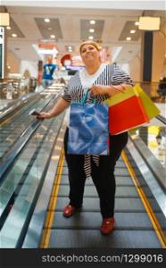 Fat woman with shopping bags on the escalator in mall. Overweight female person in supermarket, fatty shopaholic, obesity problem