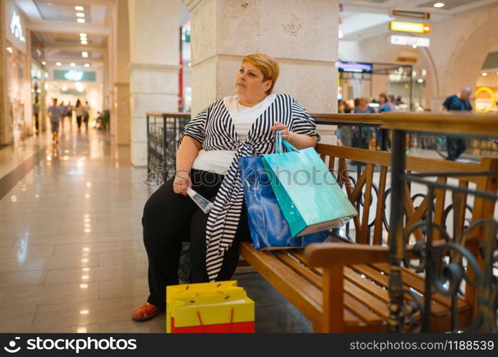 Fat woman with shopping bags sitting on the bench in mall. Overweight female person in supermarket, obesity problem