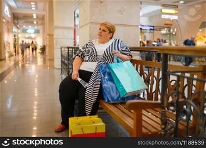 Fat woman with shopping bags sitting on the bench in mall. Overweight female person in supermarket, obesity problem