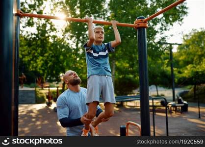 Father and boy, exercise on horizontal bar, sport training on playground outdoors. The family leads a healthy lifestyle, fitness workout in summer park. Father and boy, exercise on horizontal bar