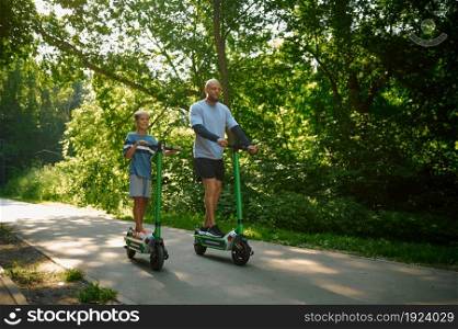 Father and boy riding on kick scooters in summer park. The family leads a healthy lifestyle, active sport workout outdoors, happy childhood. Father and boy riding on kick scooters in park