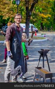 Father and child are engaged in craft. A blacksmith forges a metal blank on the anvil at a fair in the presence of spectators. Close-up.. A blacksmith forges a metal blank on the anvil at a fair in the presence of spectators.