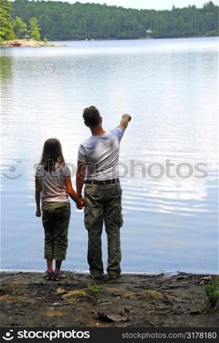 Father and daughter standing on the lake shore and looking at calm water, father pointing at somthing on the other shore